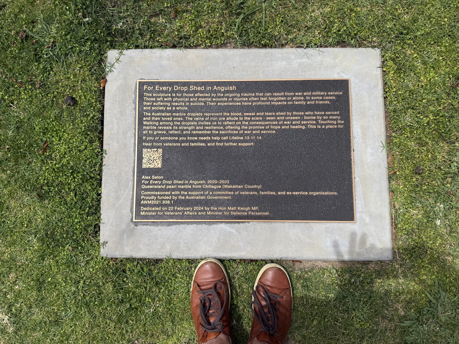 Brown leather shoes standing on grass at the foot of a bronze plaque for For Every Drop Shed in Anguish.